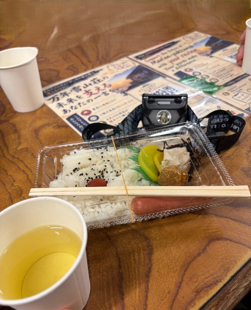 万年雪山荘の朝ご飯弁当。温かい緑茶とごはん、ソーセージ、梅干し、シューマイ、ミートボール、昆布、たくあん。